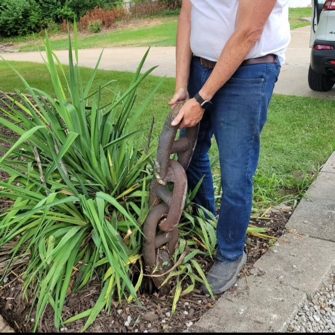 Person holding a large rusted chain near overgrown plants in a garden setting.