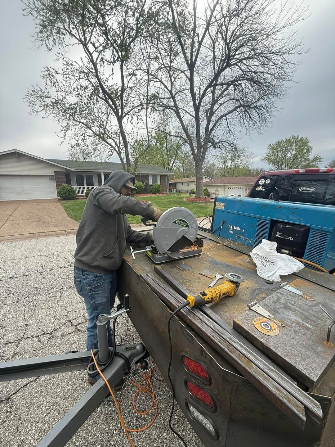 Man using a circular saw on a trailer in a residential area, surrounded by trees.