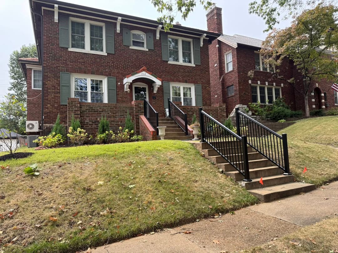Brick house with large front steps, railing, and landscaped yard. Trees in the background.