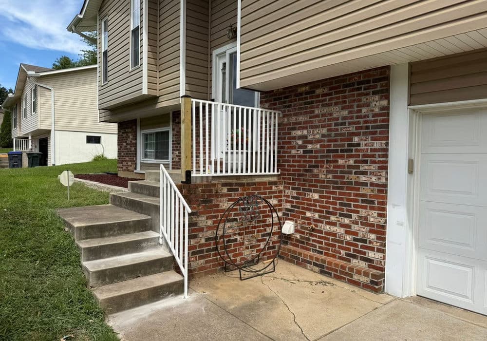 Brown brick house with steps, white railings, and a garage. Green lawn and clear blue sky.