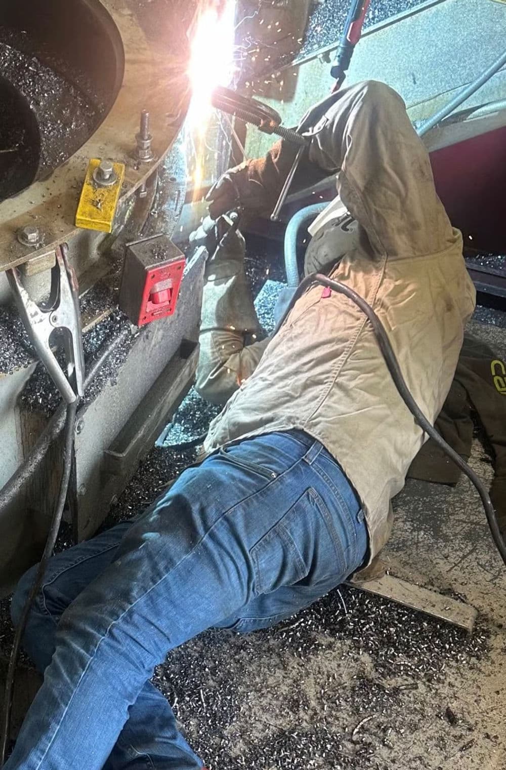 Welder working on a metal structure, sparks flying, in a workshop filled with shavings.