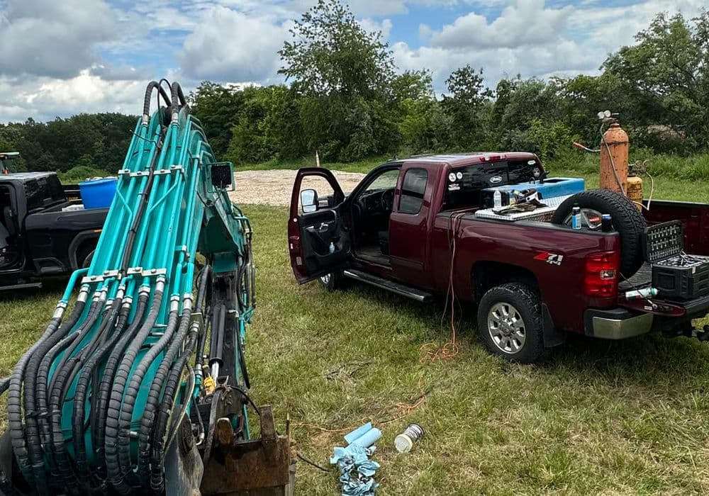 Maroon truck with equipment parked on grass, surrounded by construction tools and machinery.