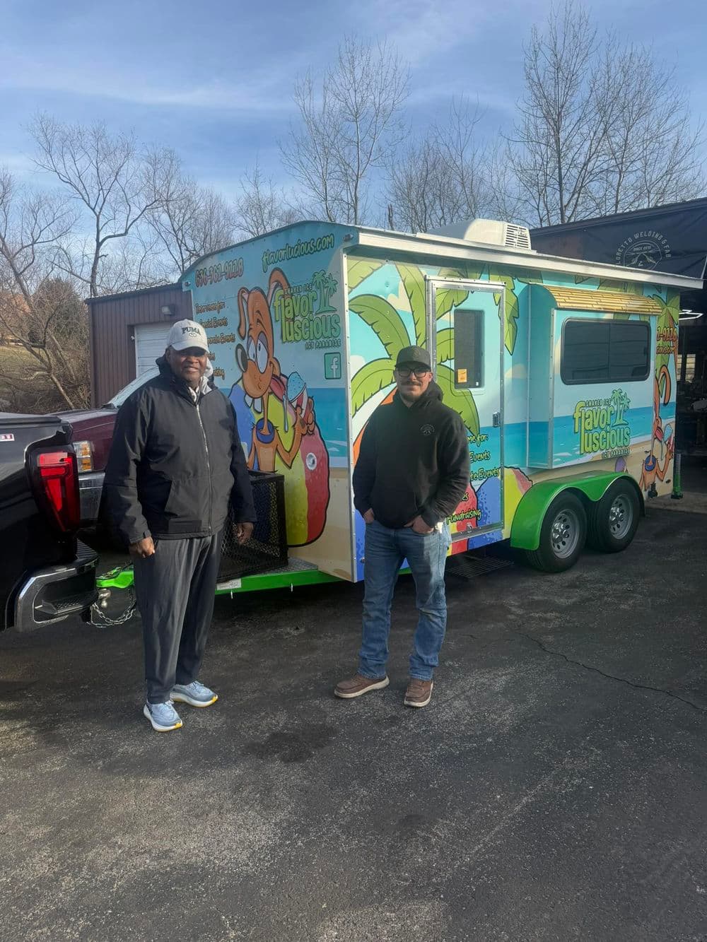 Two men stand beside a colorful food truck named "Flavor Luscious," parked outdoors.