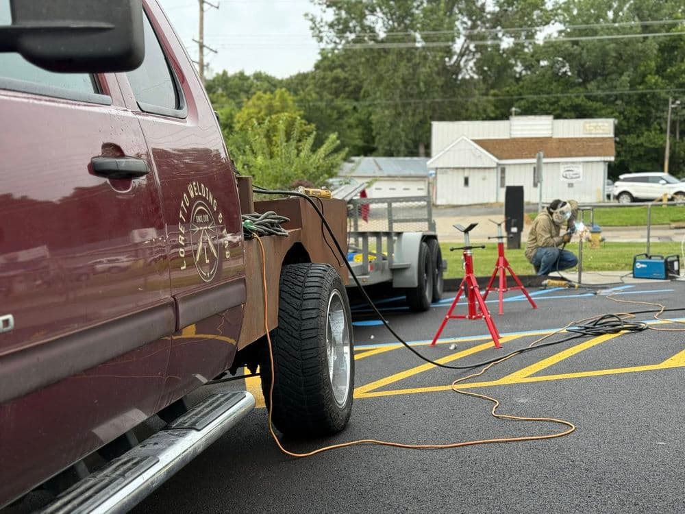 Pickup truck with trailer in foreground; worker welding in background on a job site.
