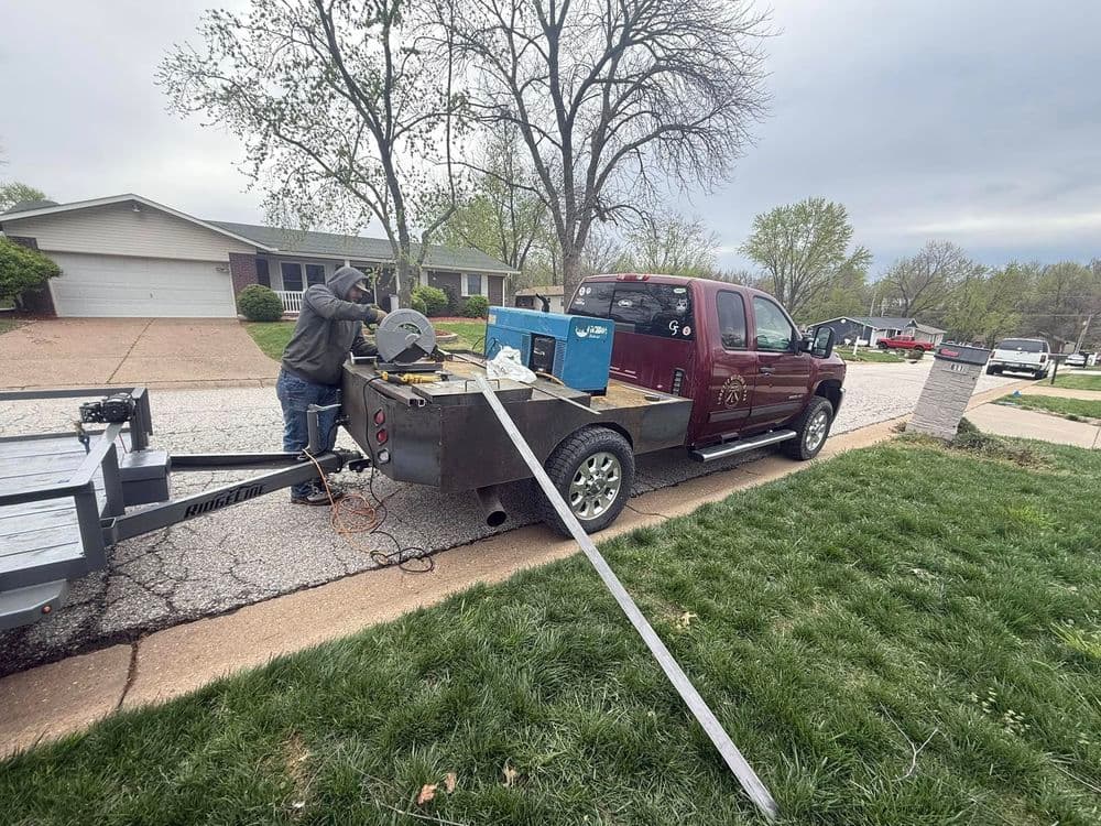 Truck with trailer and equipment parked on residential street during daytime. Worker preparing tools.