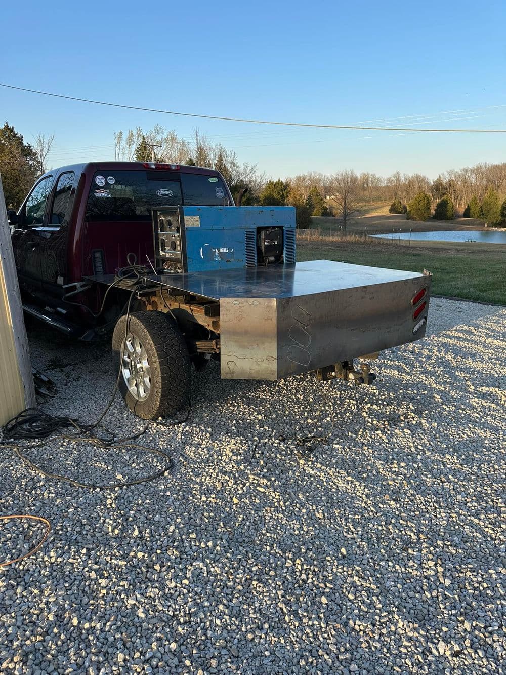 Pickup truck with a flatbed and welding equipment in a gravel driveway. Clear sky in background.