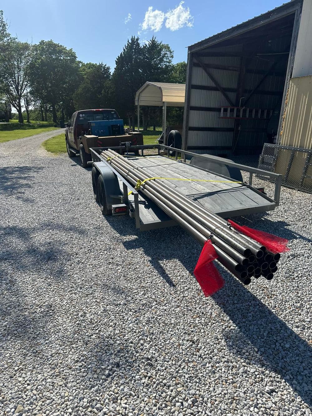 Trailer loaded with steel pipes parked on gravel driveway, surrounded by trees and a shed.