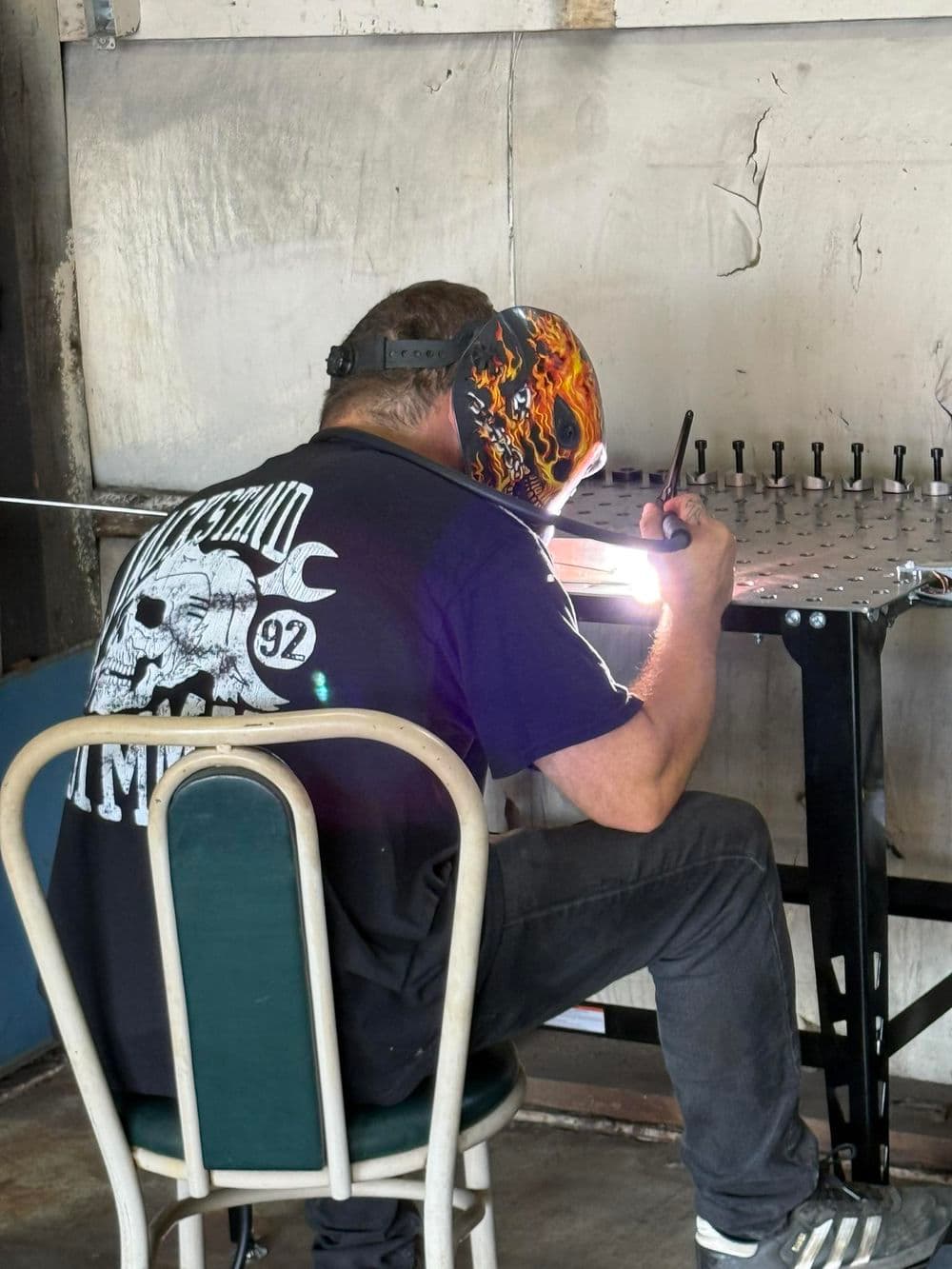 Welder in protective gear working on metal, seated on a chair in a workshop.