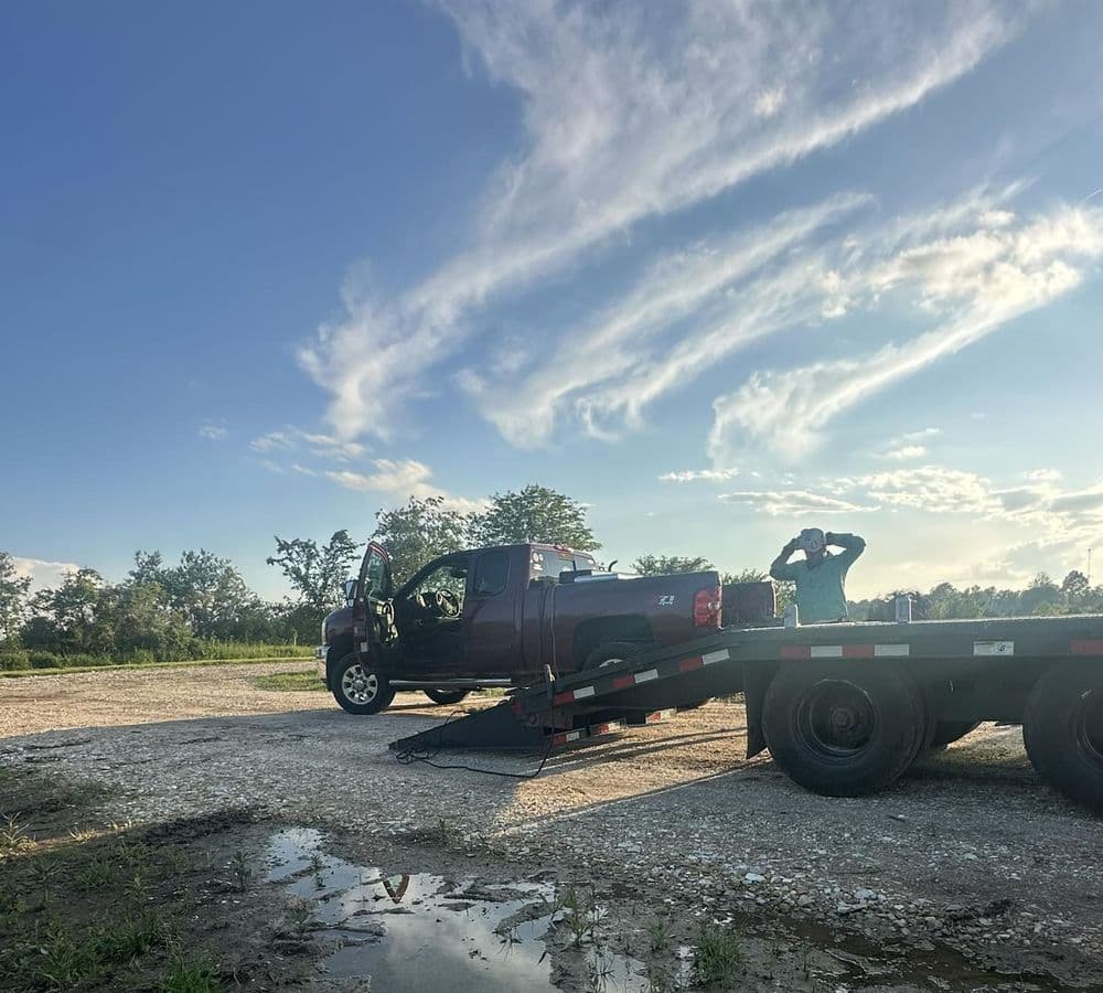Pickup truck on a trailer in an open field under a blue sky with scattered clouds.