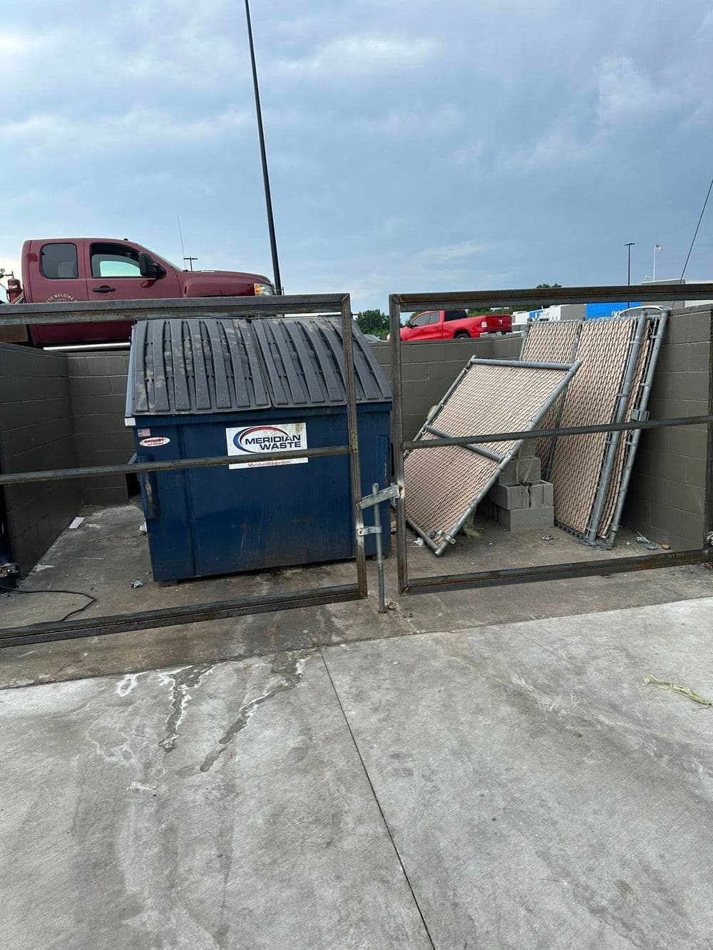 Blue dumpster behind a fence with construction materials beside it in a parking lot.