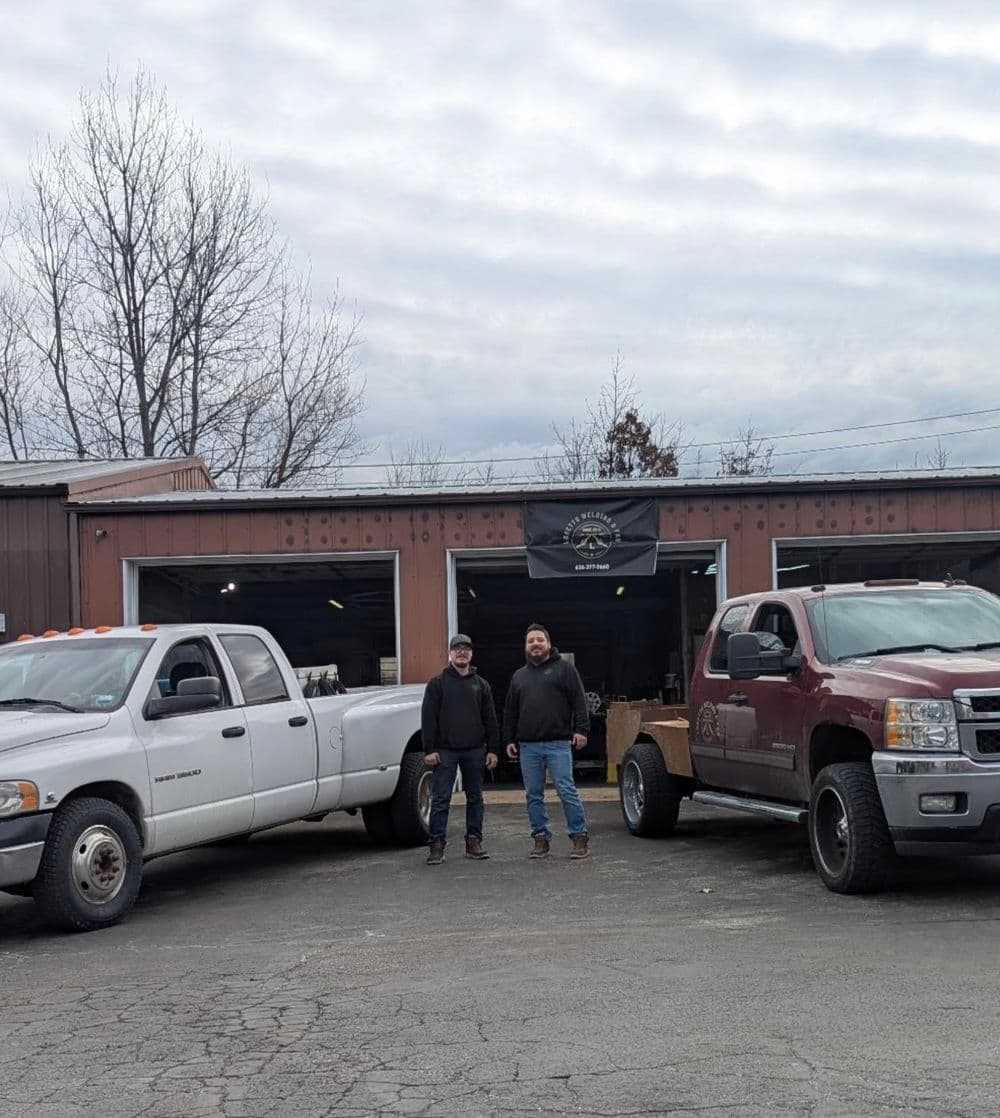 Two men standing between a white and a red pickup truck outside a workshop under a cloudy sky.